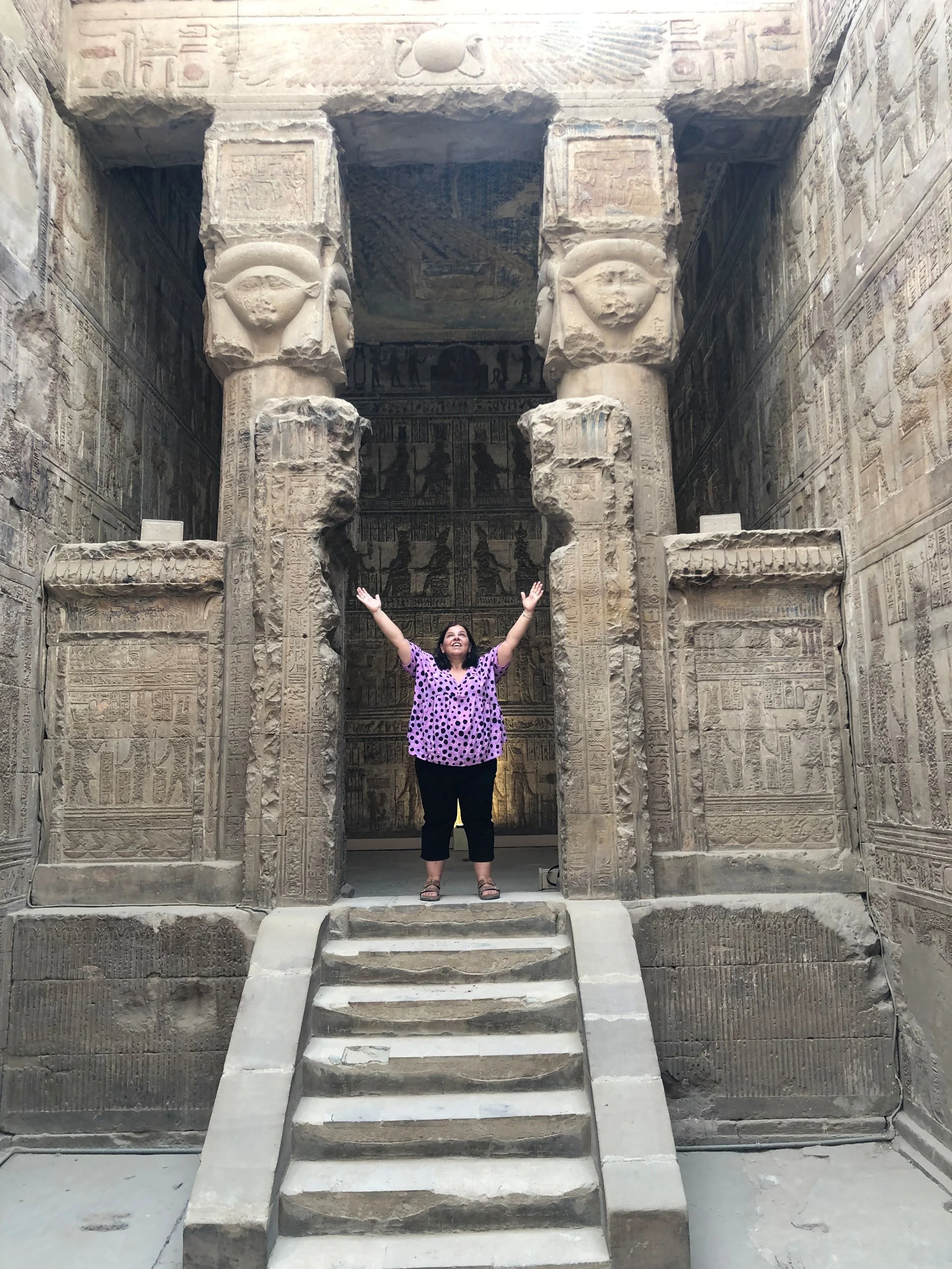 A woman standing at the entrance of an ancient Egyptian temple, raising her arms in celebration. The entrance is decorated with hieroglyphics and large stone statues of Egyptian deities.