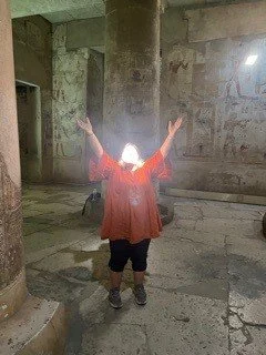 Person standing inside a large, old stone building with arms raised, facing away from the camera.