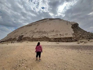 A person in a pink jacket and black pants standing in front of a large, ancient pyramid under a cloudy sky.