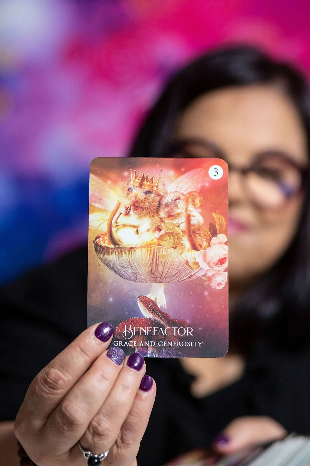 A woman holding a card with a fantasy illustration of a mouse wearing a crown and butterfly wings, sitting on a mushroom, with a snake beneath the mushroom and flowers around. The card reads "Benefactor" and "Grace and Generosity".