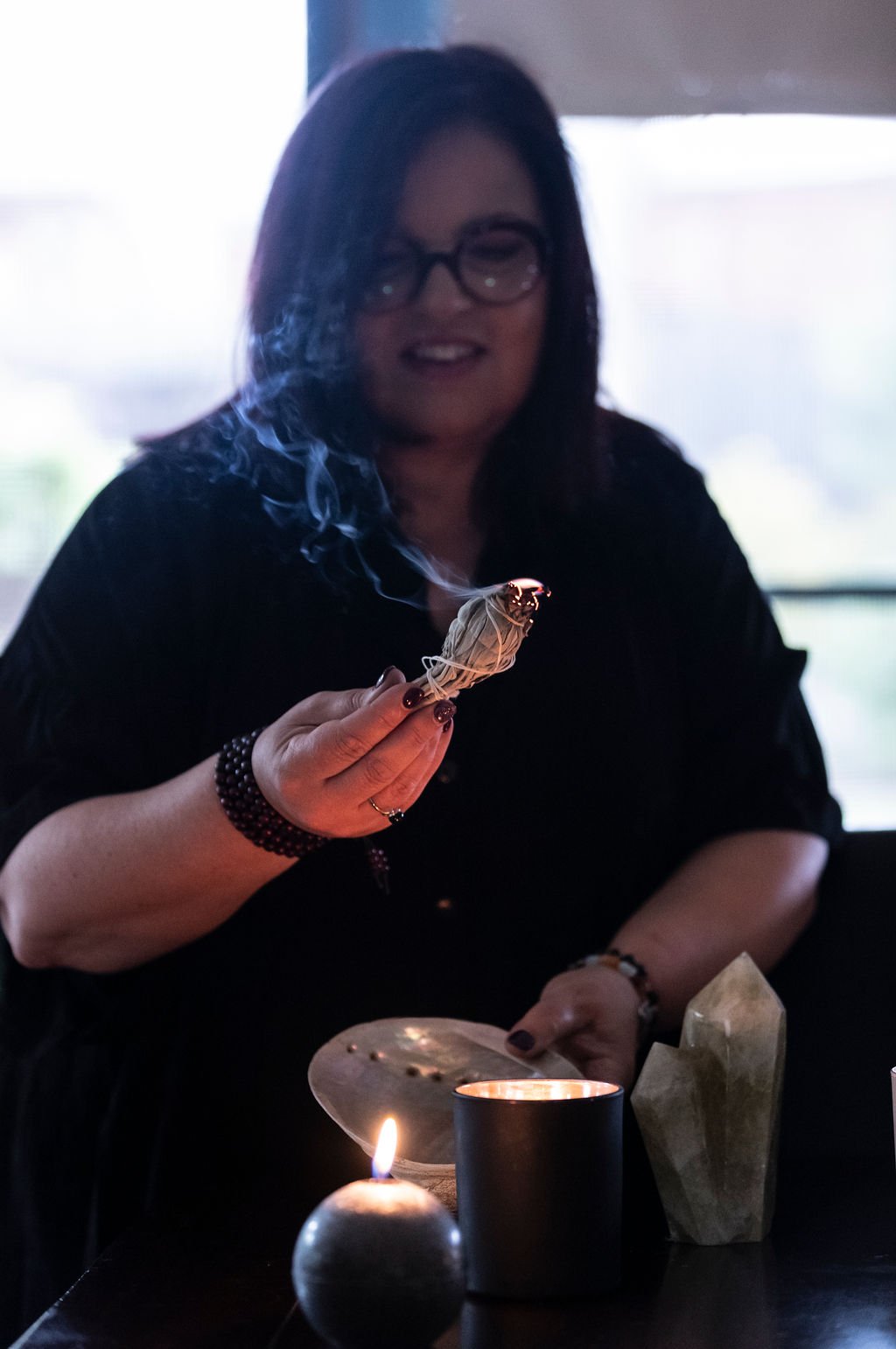 A woman with black hair and glasses holds a lit bundle of sage in her hand, with smoke rising from it. She is sitting at a table with candles and crystals, smiling at the camera.