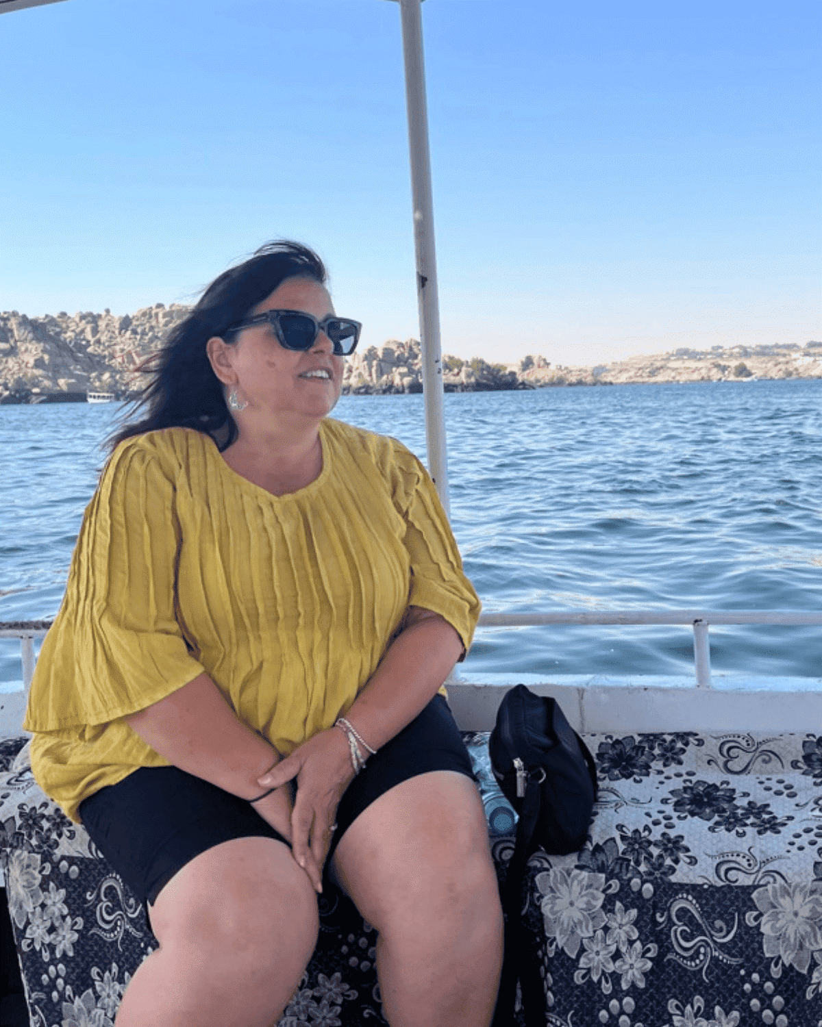 A woman in a yellow top and sunglasses sitting on a boat with water and rocky hills in the background.