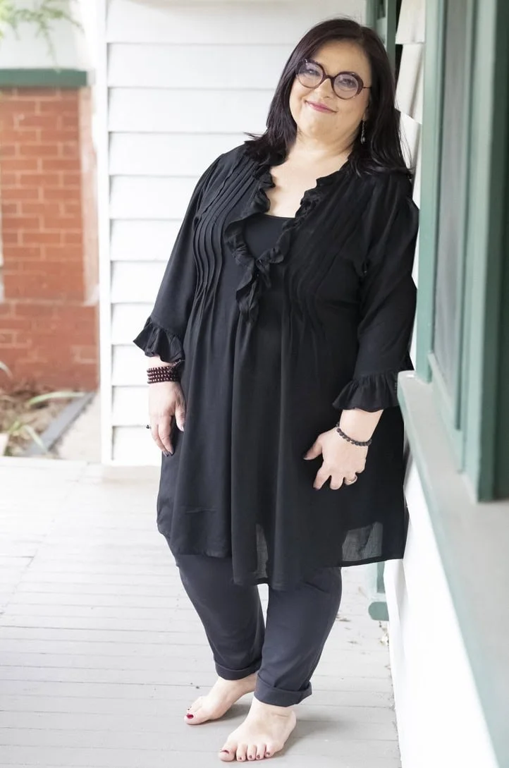 Woman in black outfit leaning against a white wall, smiling, with glasses, barefoot, on a porch.