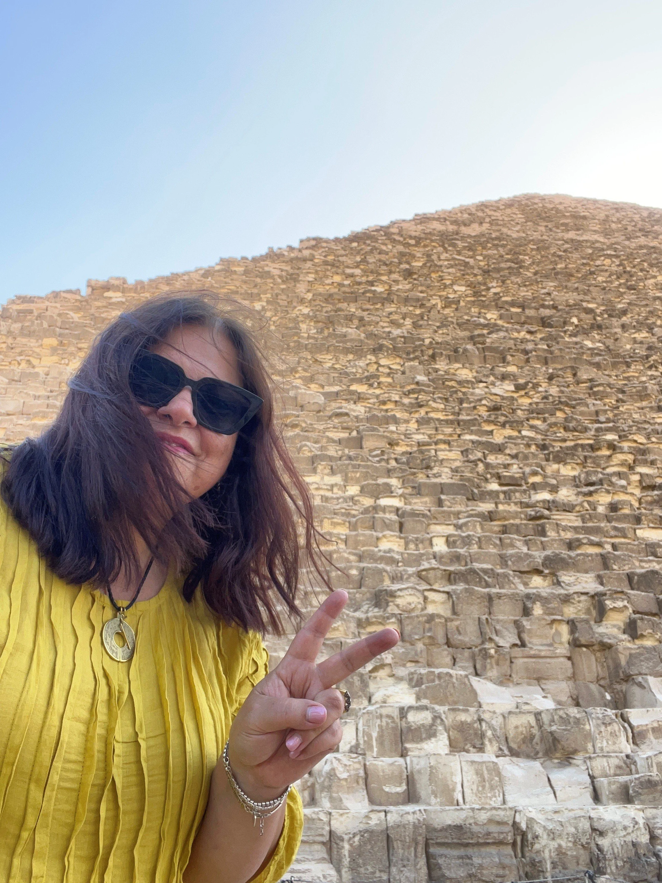 A woman with long brown hair, wearing black sunglasses, a yellow shirt, and jewelry, making a peace sign with her right hand in front of a large ancient stone pyramid.