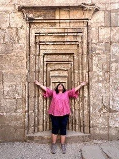 Person in a pink blouse and dark pants standing with arms outstretched in front of an ancient stone doorway with multiple rectangular frames.