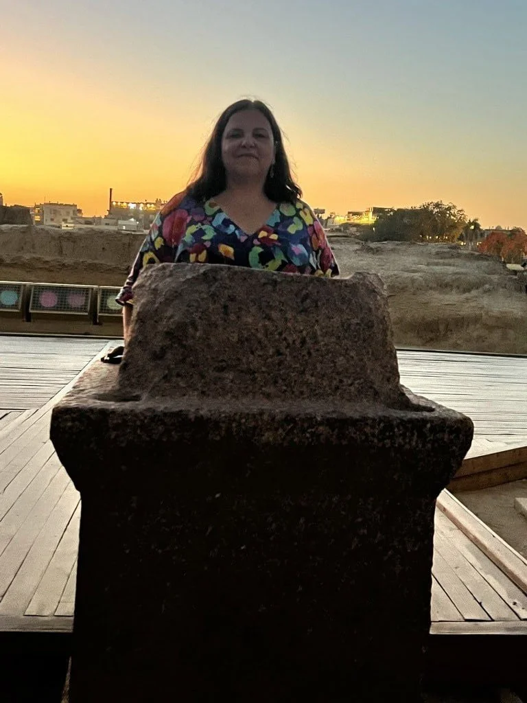A woman wearing a colorful patterned shirt standing behind an ancient stone sculpture during sunset with a desert landscape and buildings in the background.
