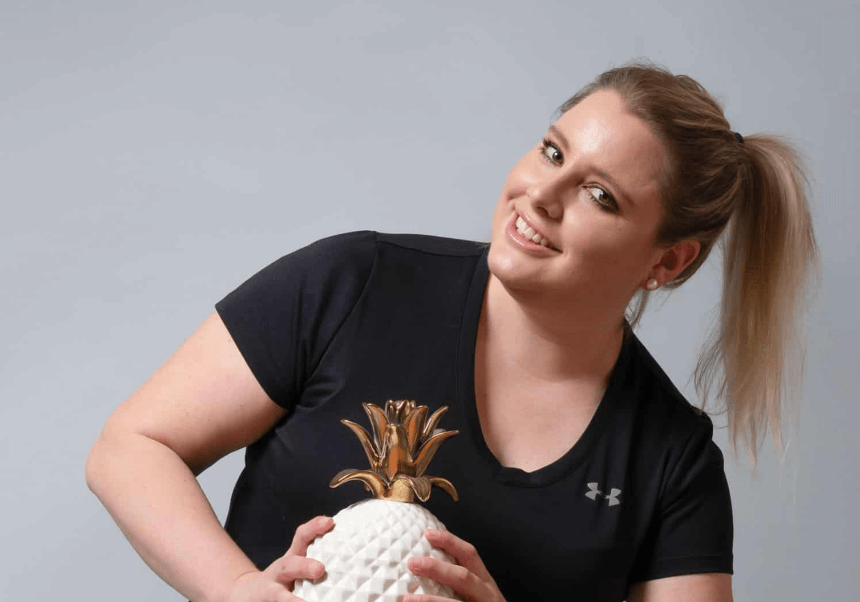 Junge Frau mit Pferdeschwanz, lächelnd, trägt ein schwarzes Under Armour T-Shirt und hält eine goldene Ananas-Trophäe.