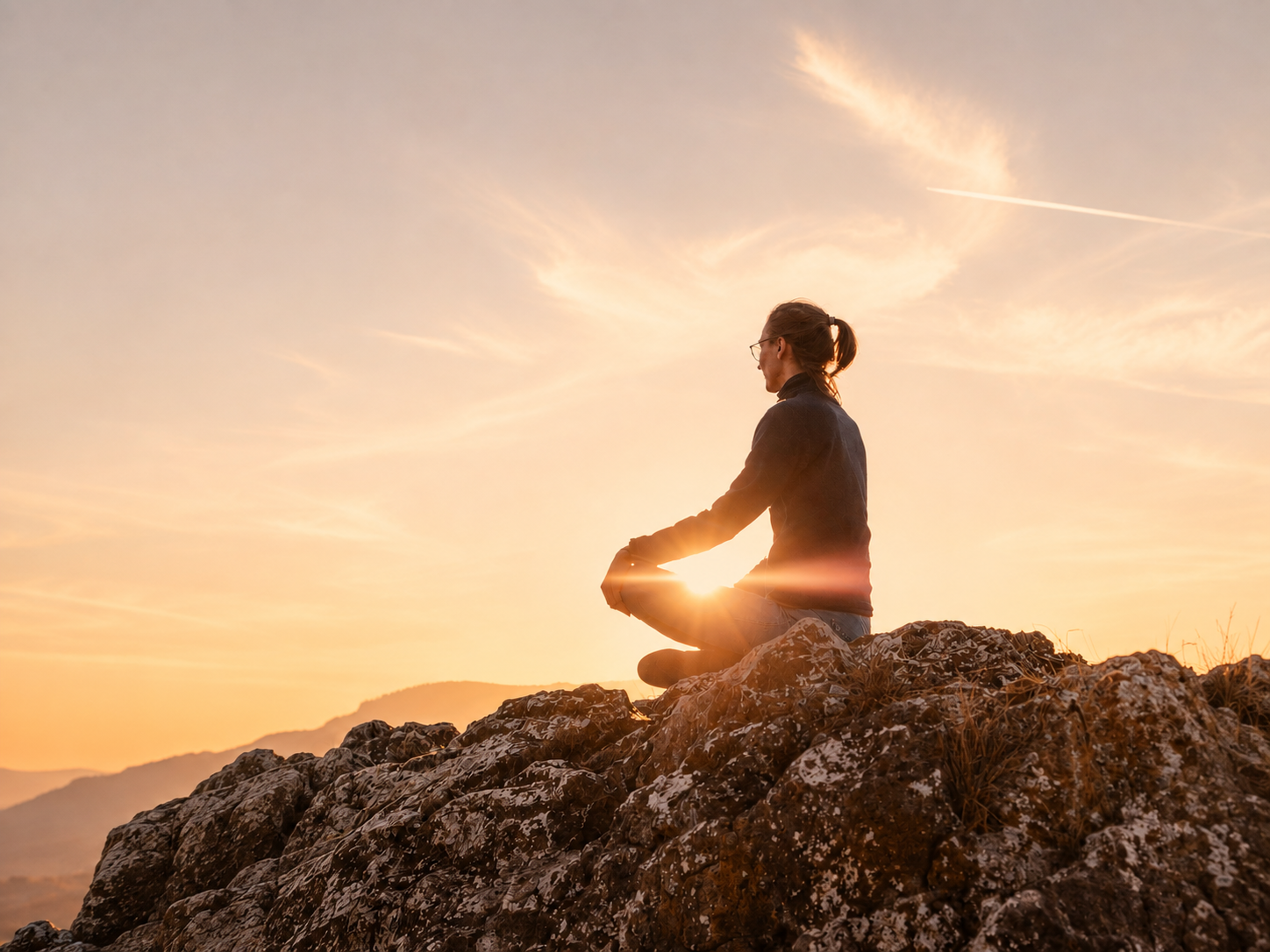 Eine Frau sitzt im Lotussitz auf einem Felsen während eines Sonnenuntergangs, die Sonne schimmert zwischen ihren Händen.