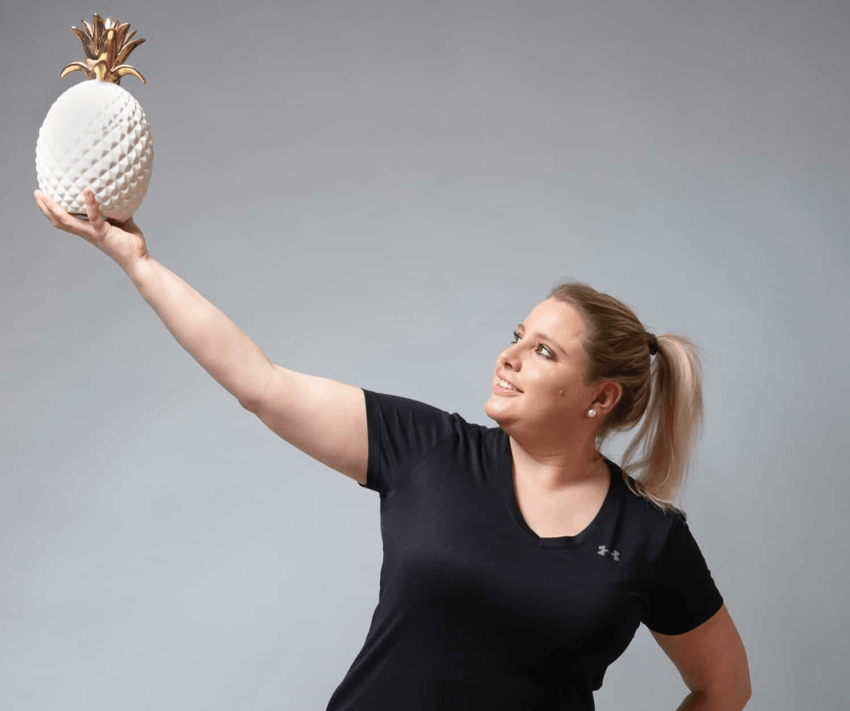 junge, lächelnde Frau mit schwarzem T-Shirt hebt eine gold-weisse Ananas-Figur hoch