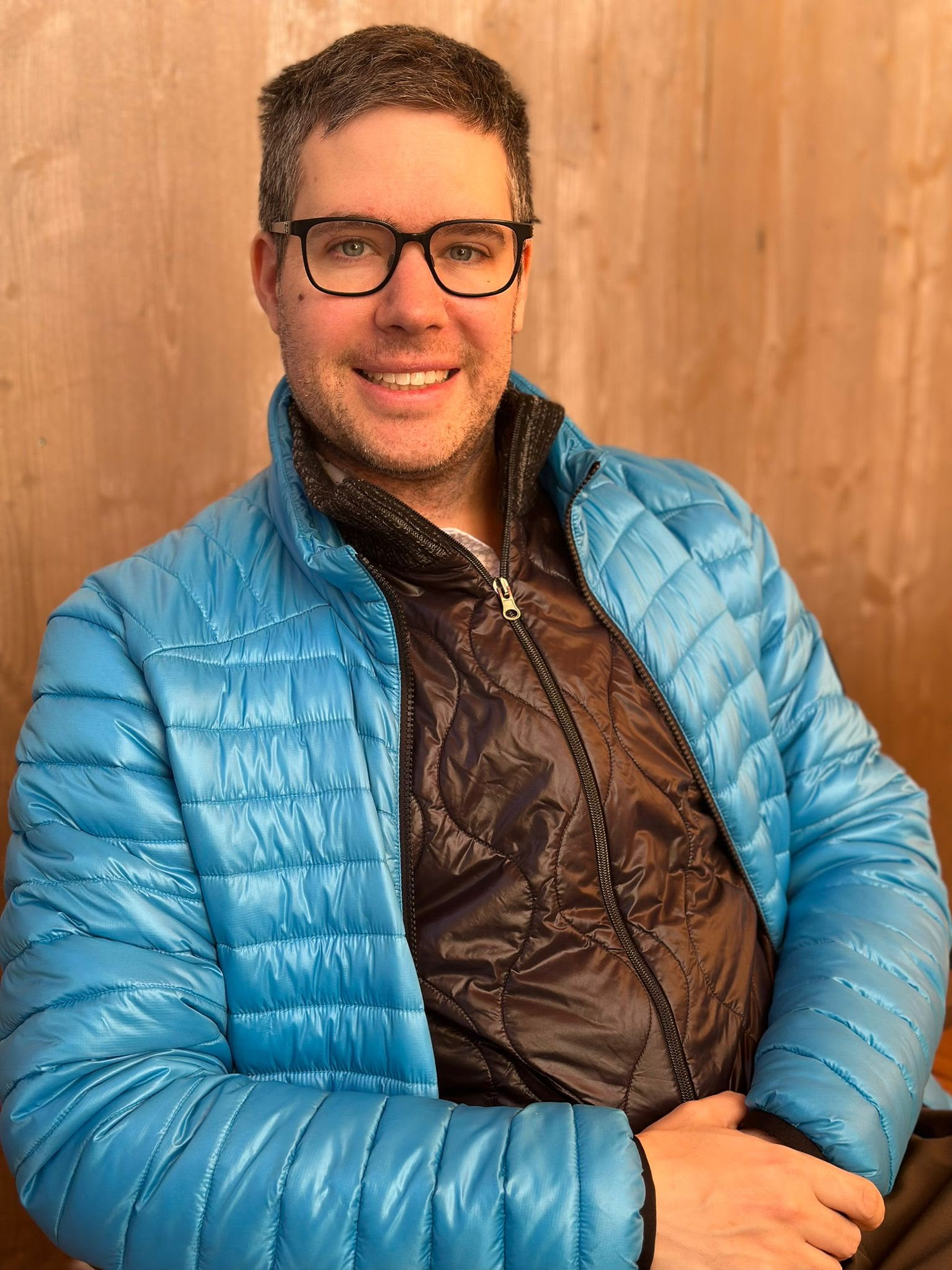 Ein junger Mann mit dunklem Haar, Brille und einem freundlichen Lächeln sitzt vor einer Holzwand und trägt eine blaue Jacke.