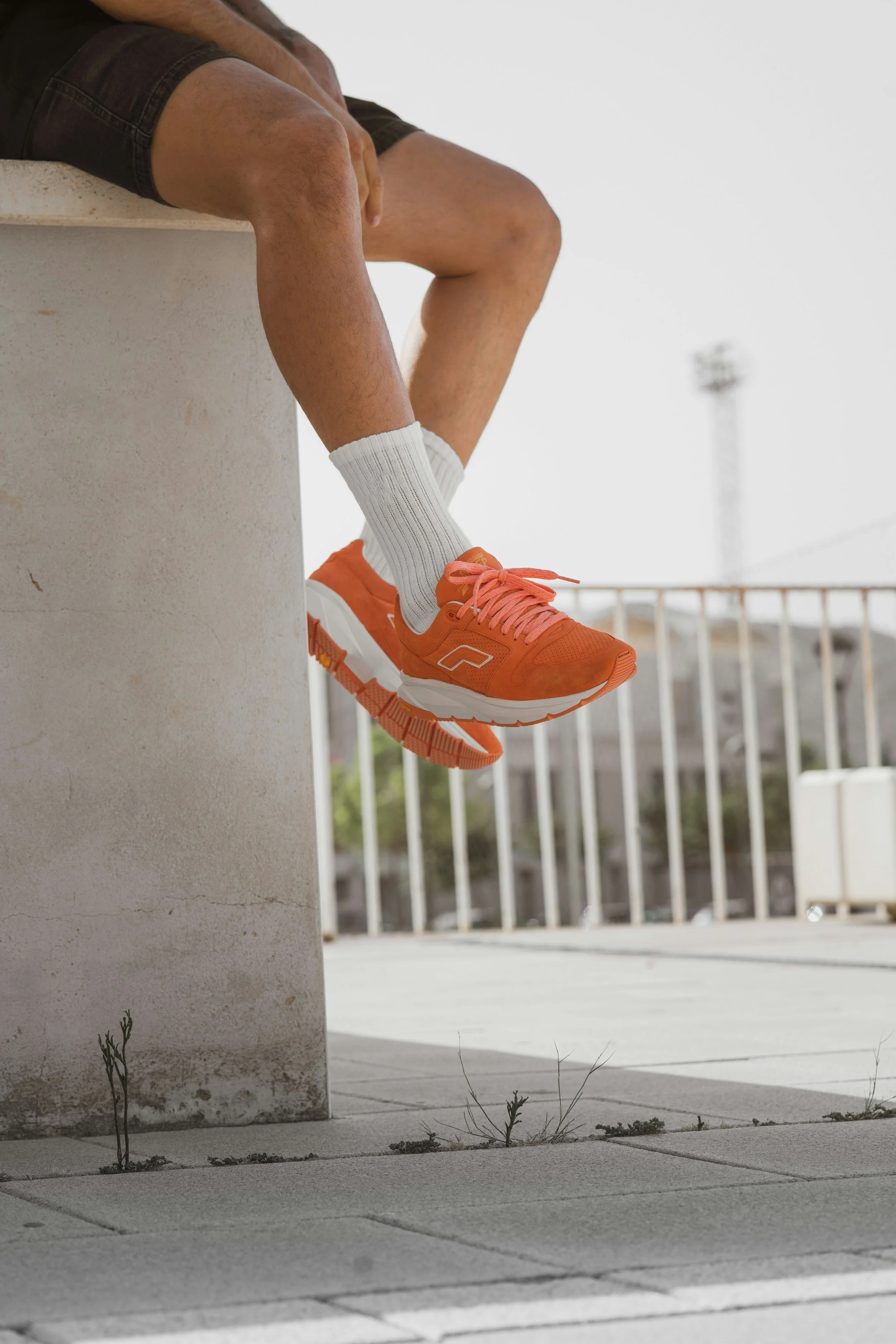 Nahaufnahme von einem Mann, der auf einer Betonmauer sitzt, mit nacktem Oberkörper und einer weißen Socke an einem Fuß, trägt orange Sportschuhe. Im Hintergrund ist ein Außenbereich mit einer Metallzaun und einem hellen Himmel zu sehen.