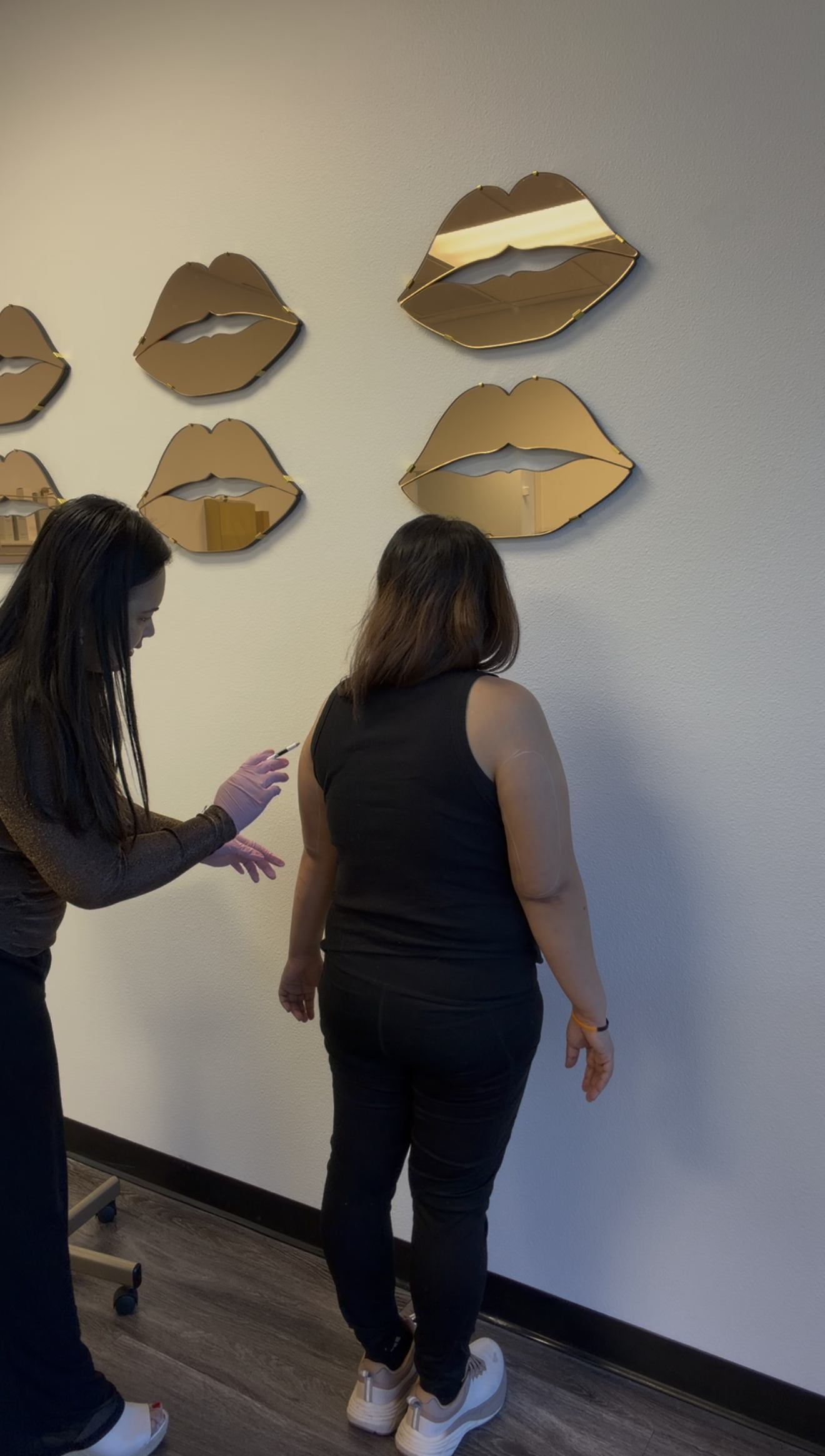 Two women, one with black hair and the other with brown hair, are standing indoors near a wall with decorative mirrors shaped like lips. The woman with brown hair is wearing a black sleeveless top, black pants, and white platform sneakers. The woman with black hair is wearing a black long-sleeve shirt, black pants, and white open-toe slide sandals, and appears to be preparing to give an injection into the other woman's arm.