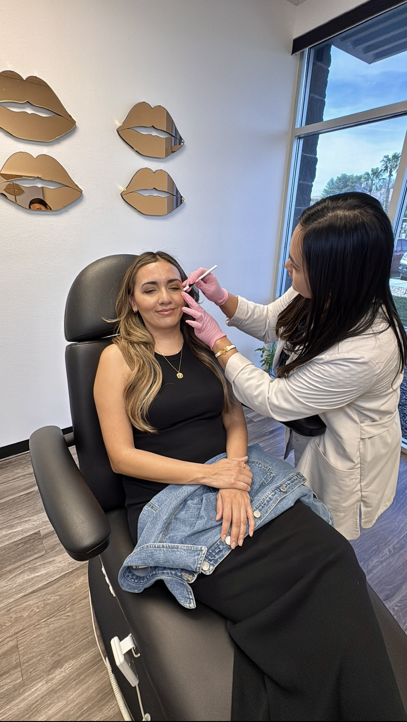 A woman receiving a cosmetic treatment on her face by a professional in a modern clinic with decorative mirrors on the wall and a large window in the background.
