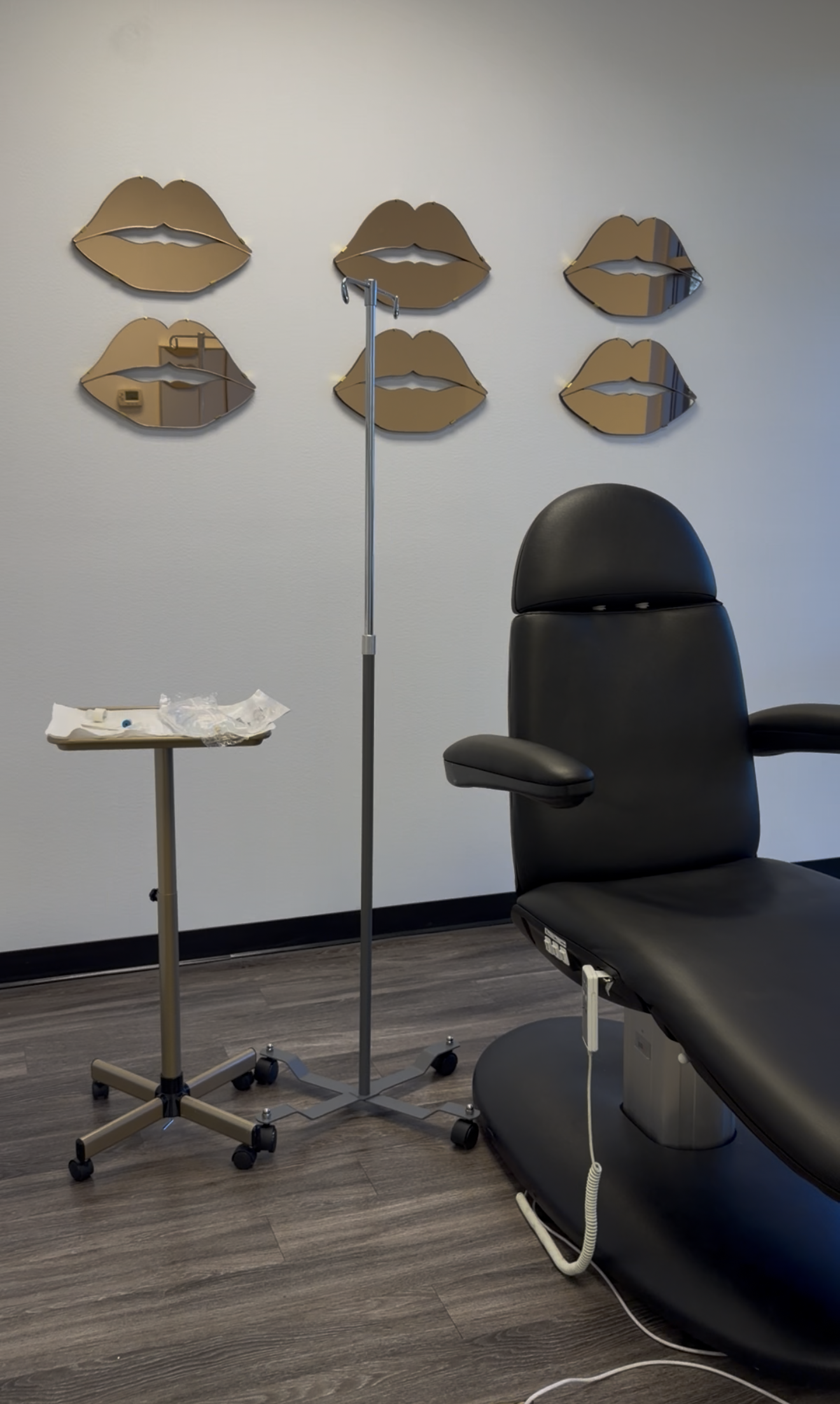 A medical examination room with a black examination chair, a IV stand, and a small medical tray. Six decorative mirrors in the shape of lips are mounted on a gray wall.