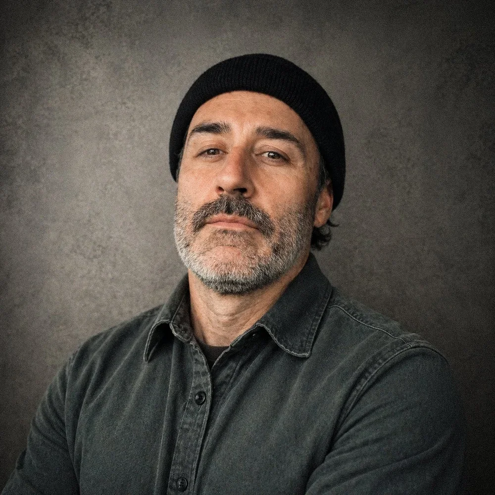 A middle-aged man with a beard and mustache, wearing a black beanie and a dark denim shirt, standing against a dark textured background.