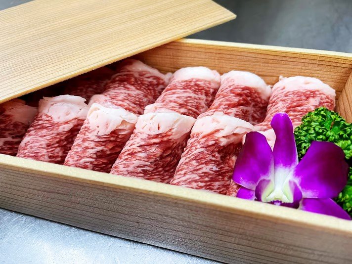 木の箱に入った薄切りの牛肉と緑の野菜と紫の花
