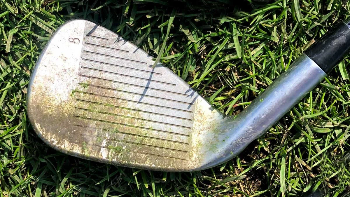 A golf club lying on the grass, showing a worn and dirty metal head with engraved lines.