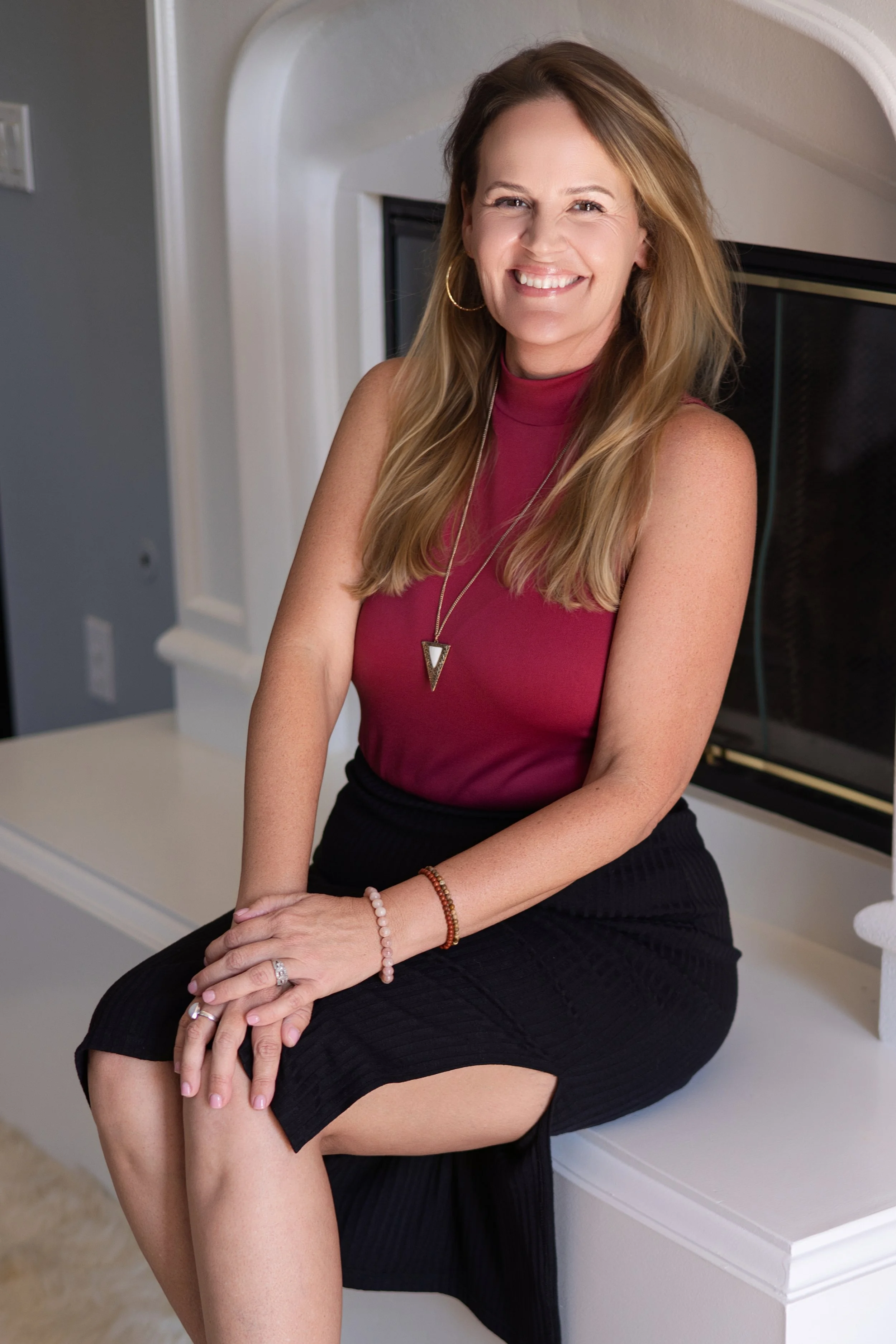 A woman with long blond hair, wearing a sleeveless burgundy top and black skirt, sitting on a windowsill with her hands resting on her lap, smiling at the camera.