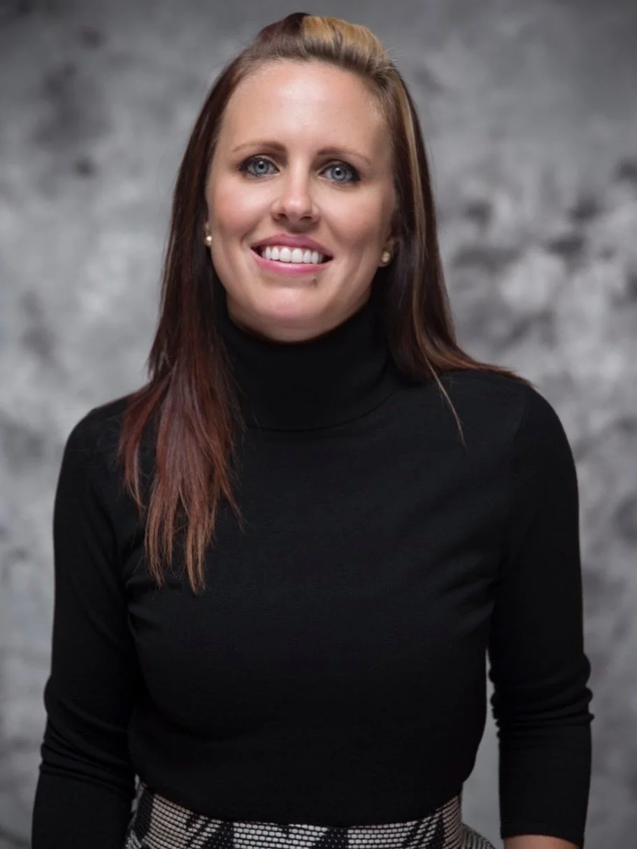 A woman with brown hair and blue eyes, smiling, wearing a black turtleneck sweater and a patterned skirt, standing in front of a gray textured background.