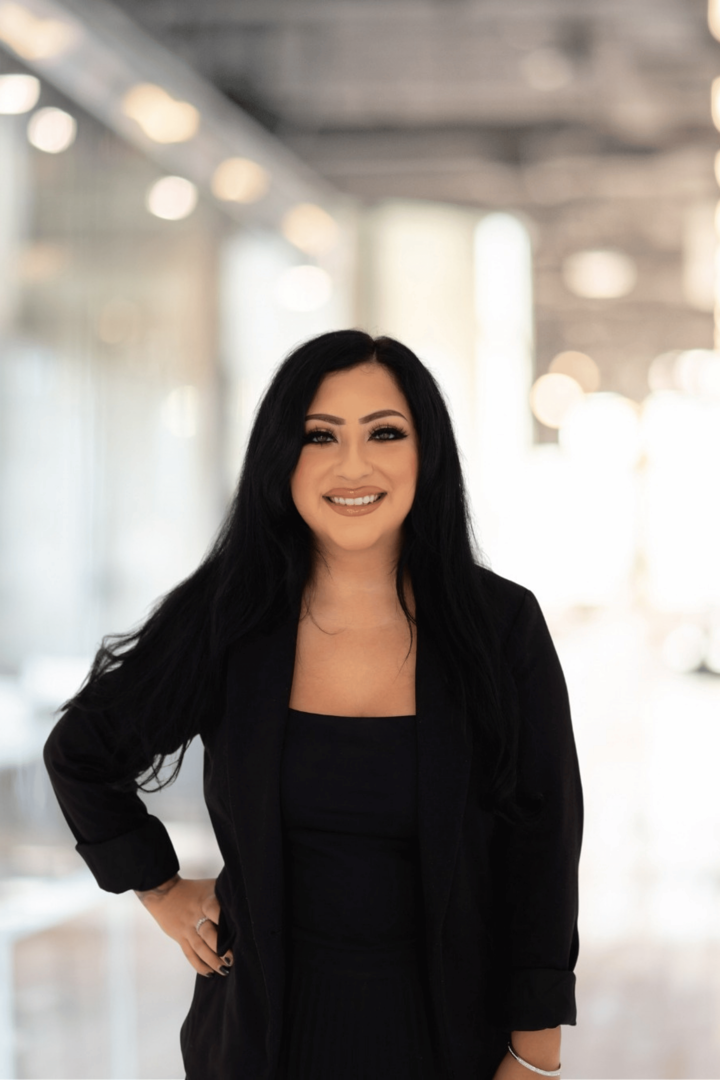 Woman with long black hair smiling, wearing a black jacket, standing in a brightly lit corridor with blurred lights in the background.
