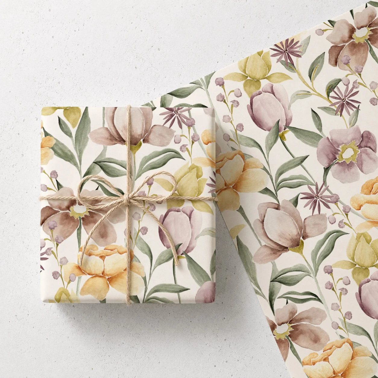 A gift wrapped in floral paper with pastel-colored flowers and green leaves, tied with a light brown twine bow, placed on white surface with additional floral wrapping paper nearby.