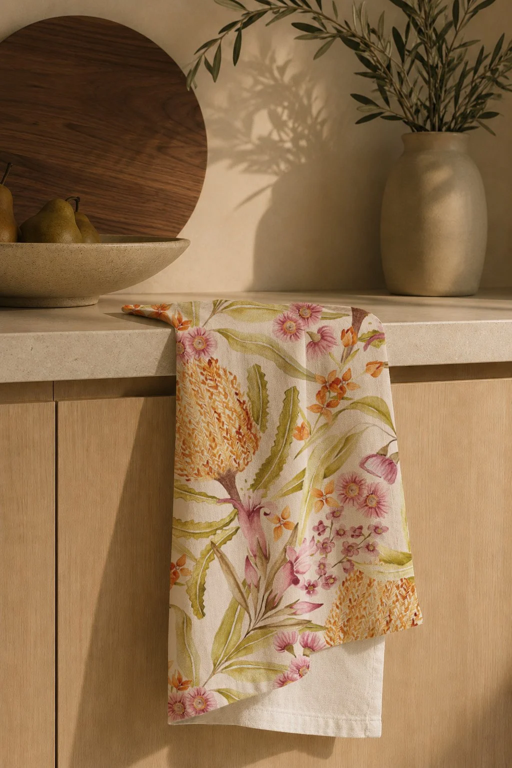 A kitchen countertop with a floral-patterned towel hanging over the edge, a beige vase with leafy branches, a decorative wooden tray, and a plate with pears.