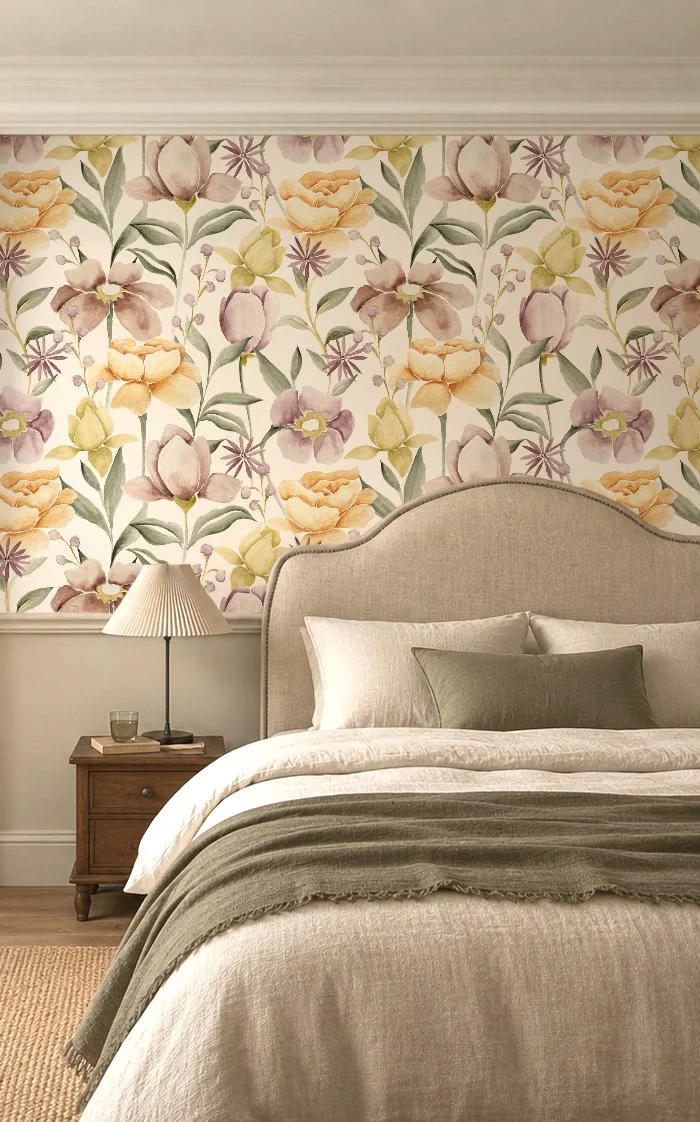 A cozy bedroom with a floral wallpaper featuring pastel-colored flowers, a beige upholstered headboard, a wooden bedside table with a lamp, glass of water, and books, and a bed with white and olive-green pillows and a beige blanket.
