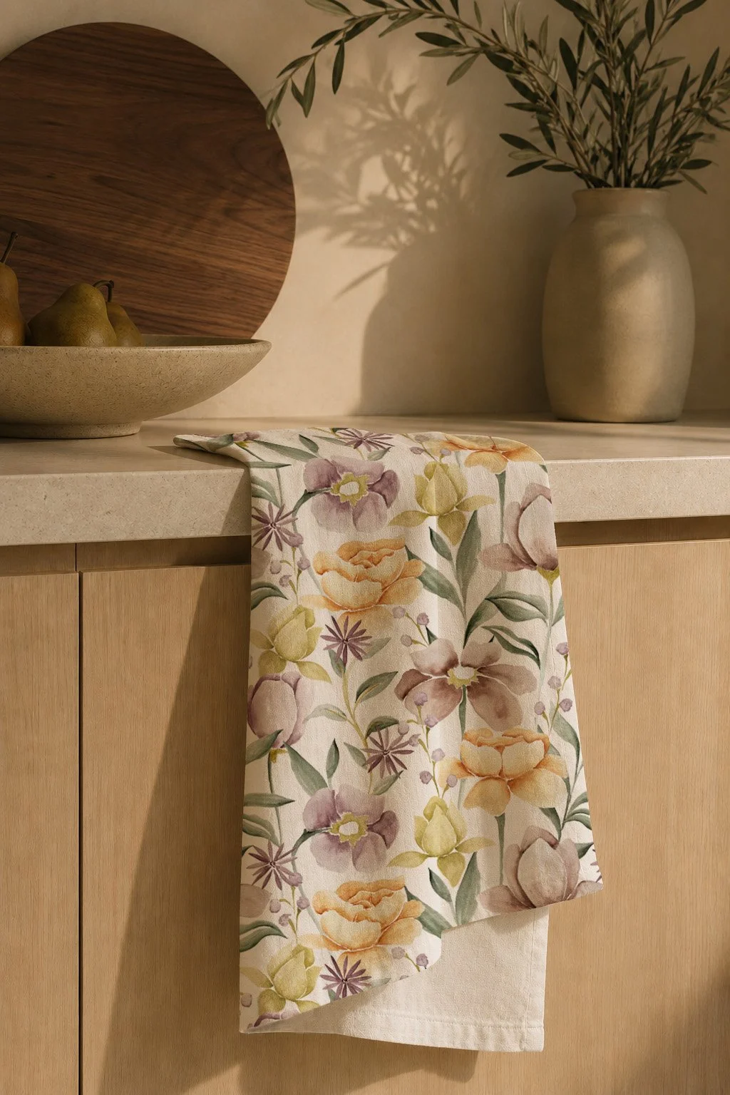 A decorative kitchen countertop with a floral-patterned cloth hanging from the edge, a beige bowl with pears, a wooden plate, and a large ceramic vase with green leafy branches casting shadows on the wall.