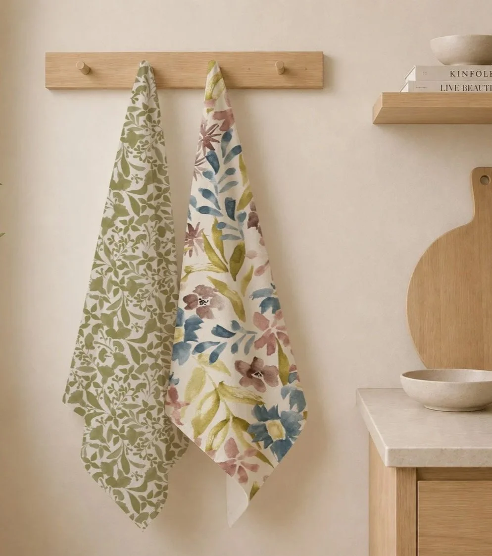 Two patterned kitchen towels hanging on a wooden wall hook in a kitchen with light-colored walls and wooden shelves.