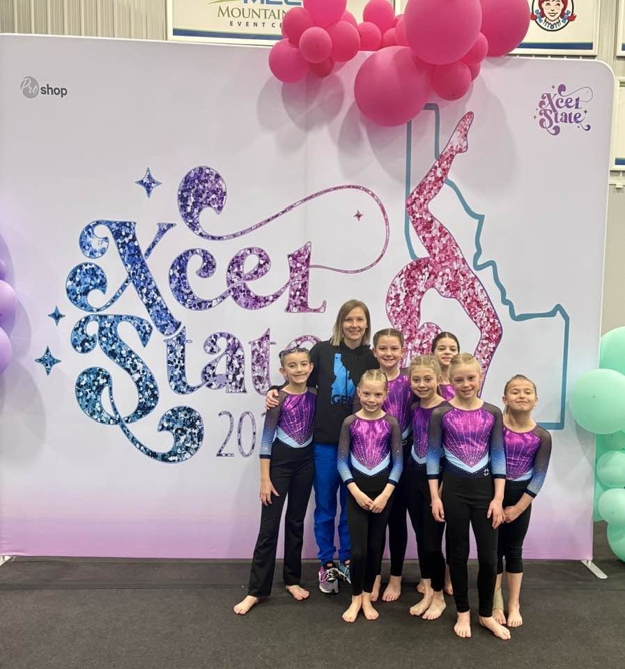 Group of young female gymnasts in matching purple and black leotards posing with their coach in front of an 'Xcel State' event backdrop decorated with balloons and colorful lettering.