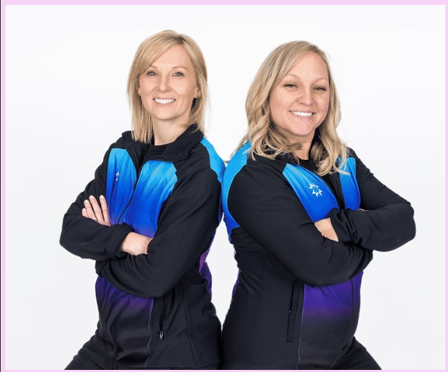Two women with blonde hair wearing black and blue jackets standing with arms crossed, smiling, against a plain white background.