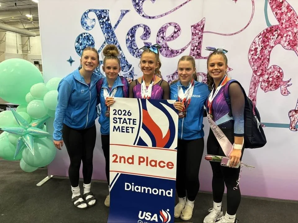 Five young female gymnasts and their coach at a gymnastics meet, holding a sign that reads '2026 State Meet 2nd Place Diamond,' with medals around their necks and a festive background with balloons and a unicorn illustration.