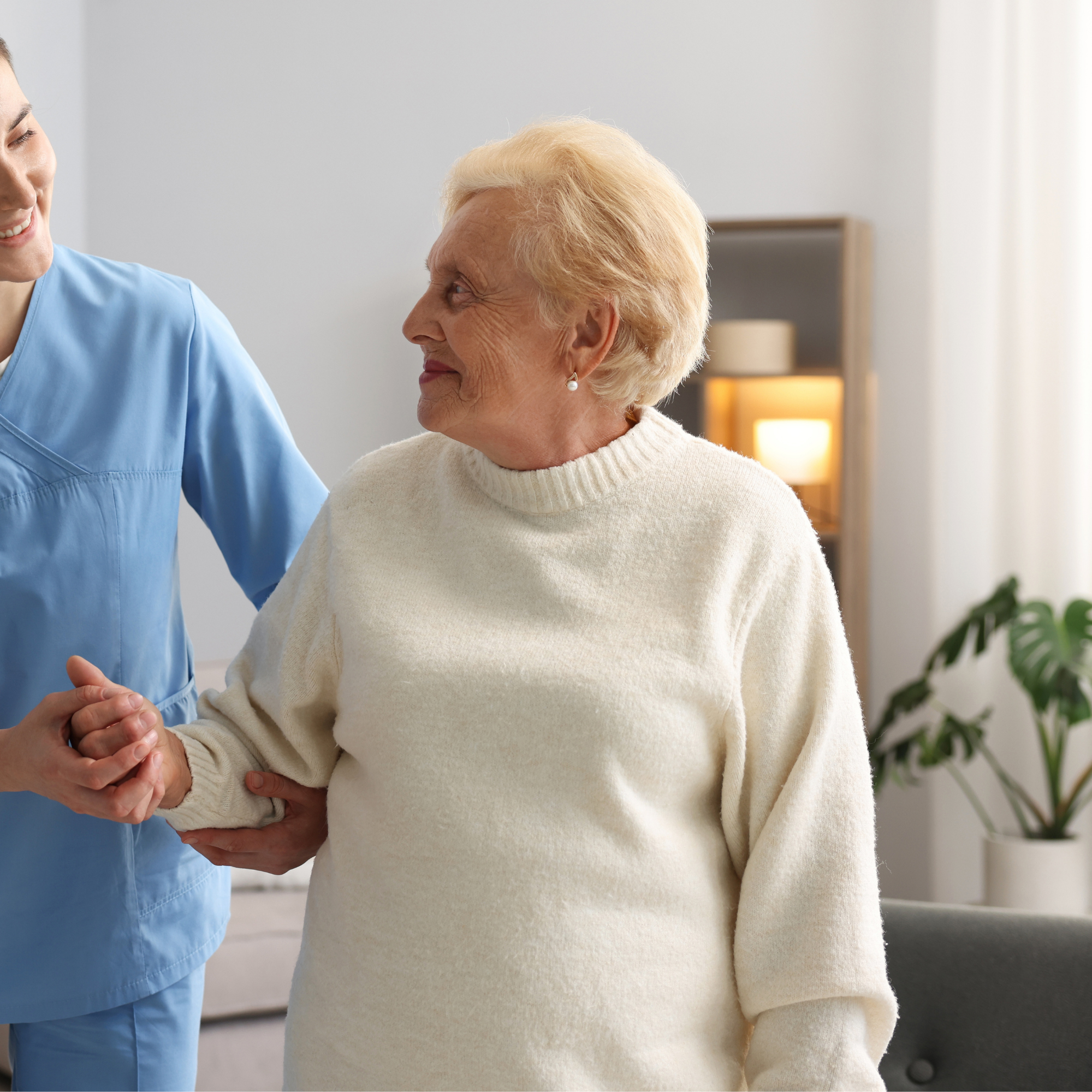 A healthcare worker in blue scrubs holding hands with an elderly woman in a cream sweater, smiling and looking at each other in a brightly lit room with a lamp and green plant in the background.