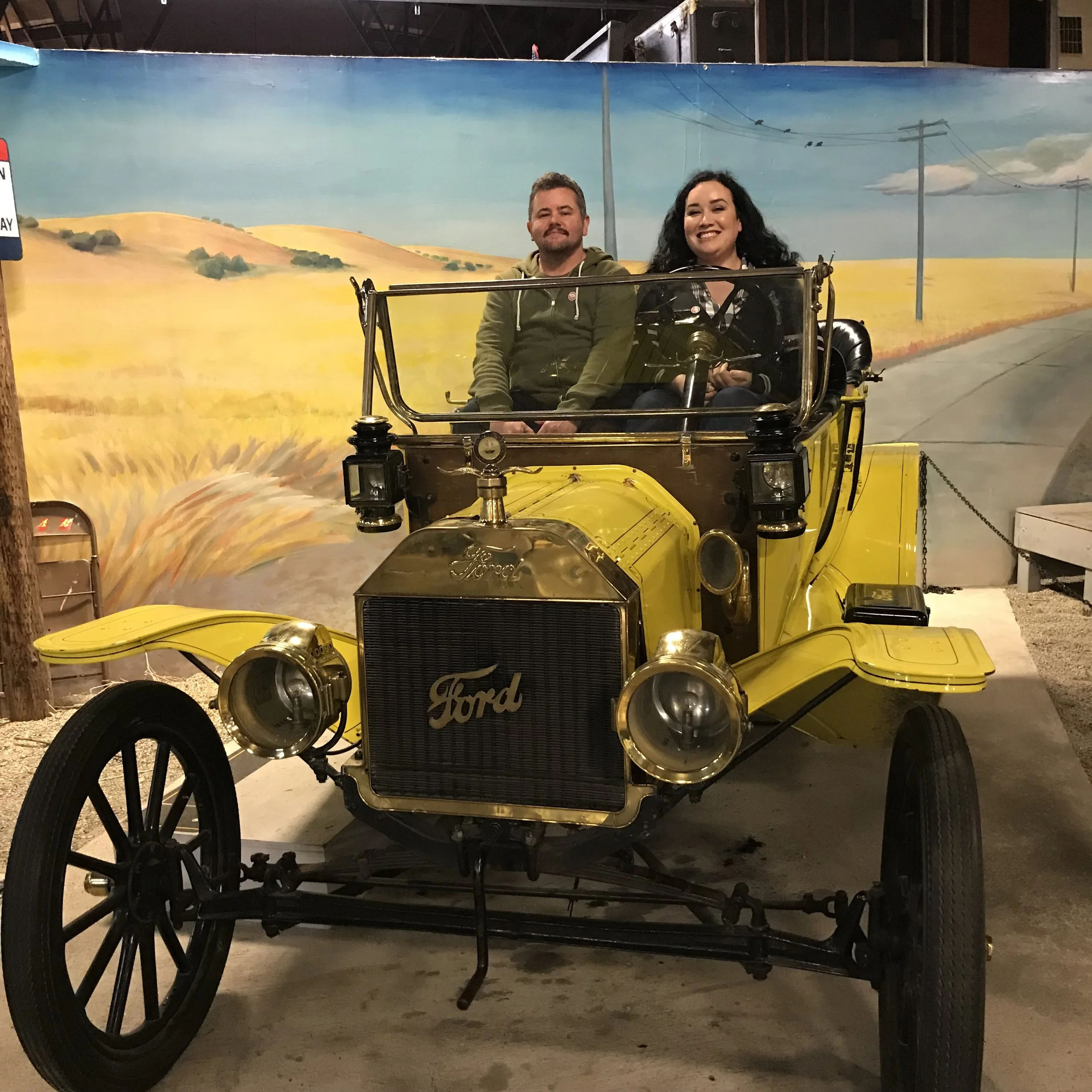 A vintage yellow Model T Ford car with black wheels and gold details, including a gold radiator, is displayed indoors. The car is in front of a painted mural of a countryside scene with rolling hills, power lines, and a blue sky with clouds. Two people are sitting in the car, one man and one woman, smiling and looking at the camera.