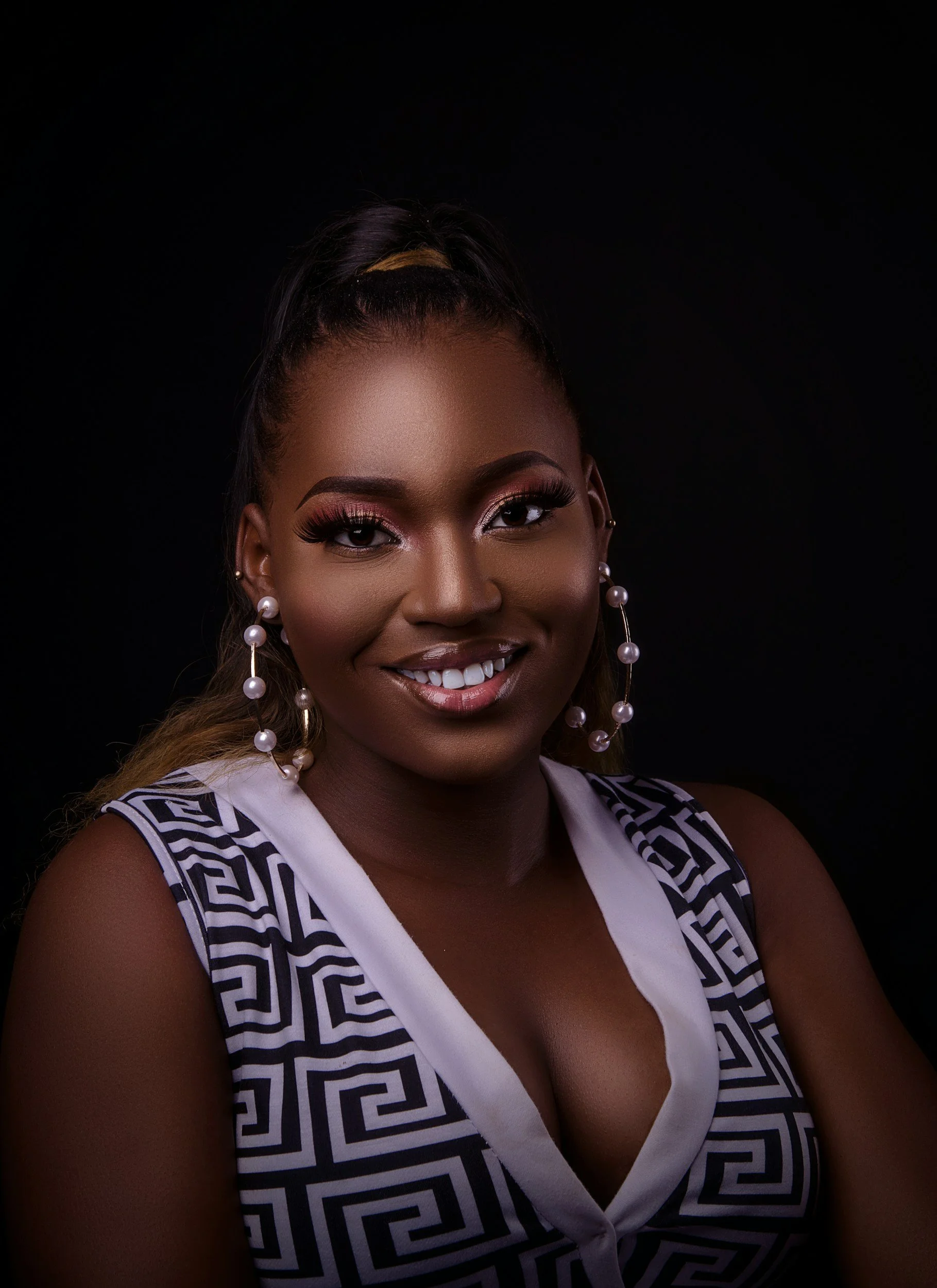 Close-up portrait of a smiling woman with braids, earrings, and a patterned dress against a black background.