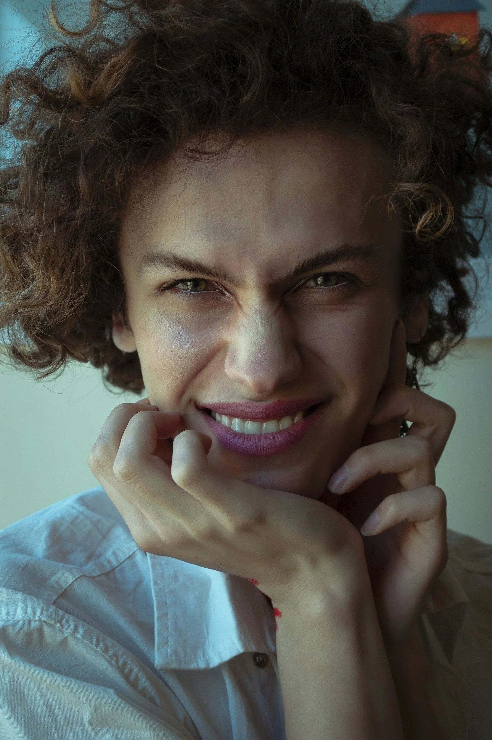 Close-up of a woman with curly hair smiling, holding her chin with both hands, wearing a light blue shirt.