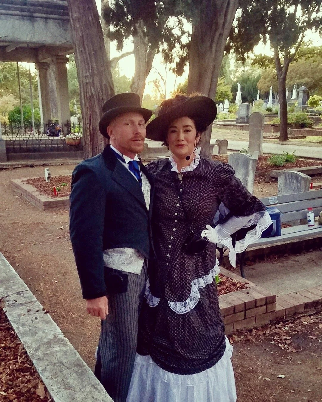 Two people dressed in Victorian or historical costumes, a man in a dark coat and tall hat, and a woman in a black dress with white lace and a wide-brimmed hat, standing outdoors in a cemetery with trees, tombstones, and a bench in the background.