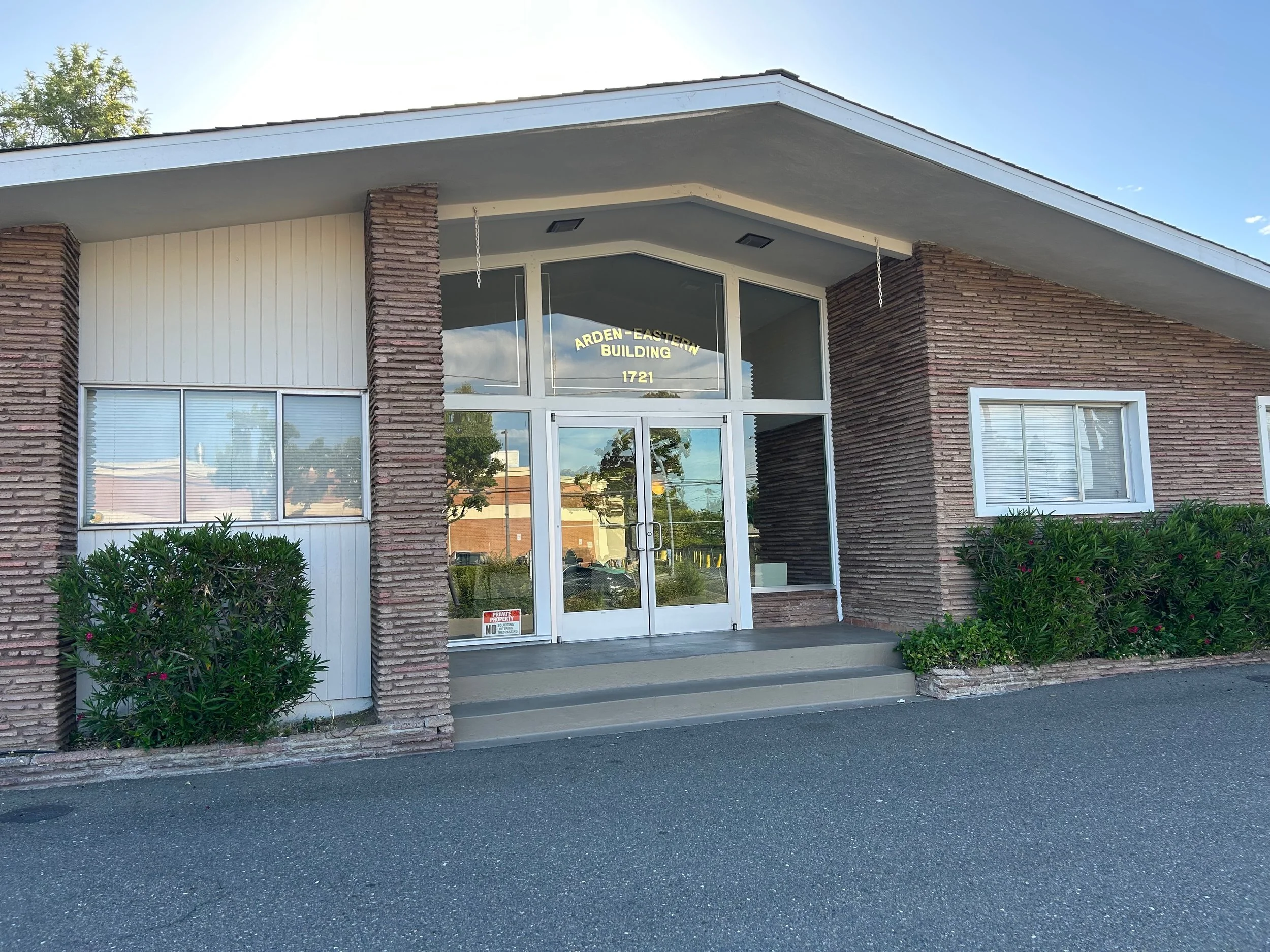 Front of a modern brick office or commercial building with large windows, a glass door, and a sign that reads 'ARDen-Eastern Building 1721'. There are bushes and shrubs in front of the building, with a sidewalk and asphalt street in the foreground.