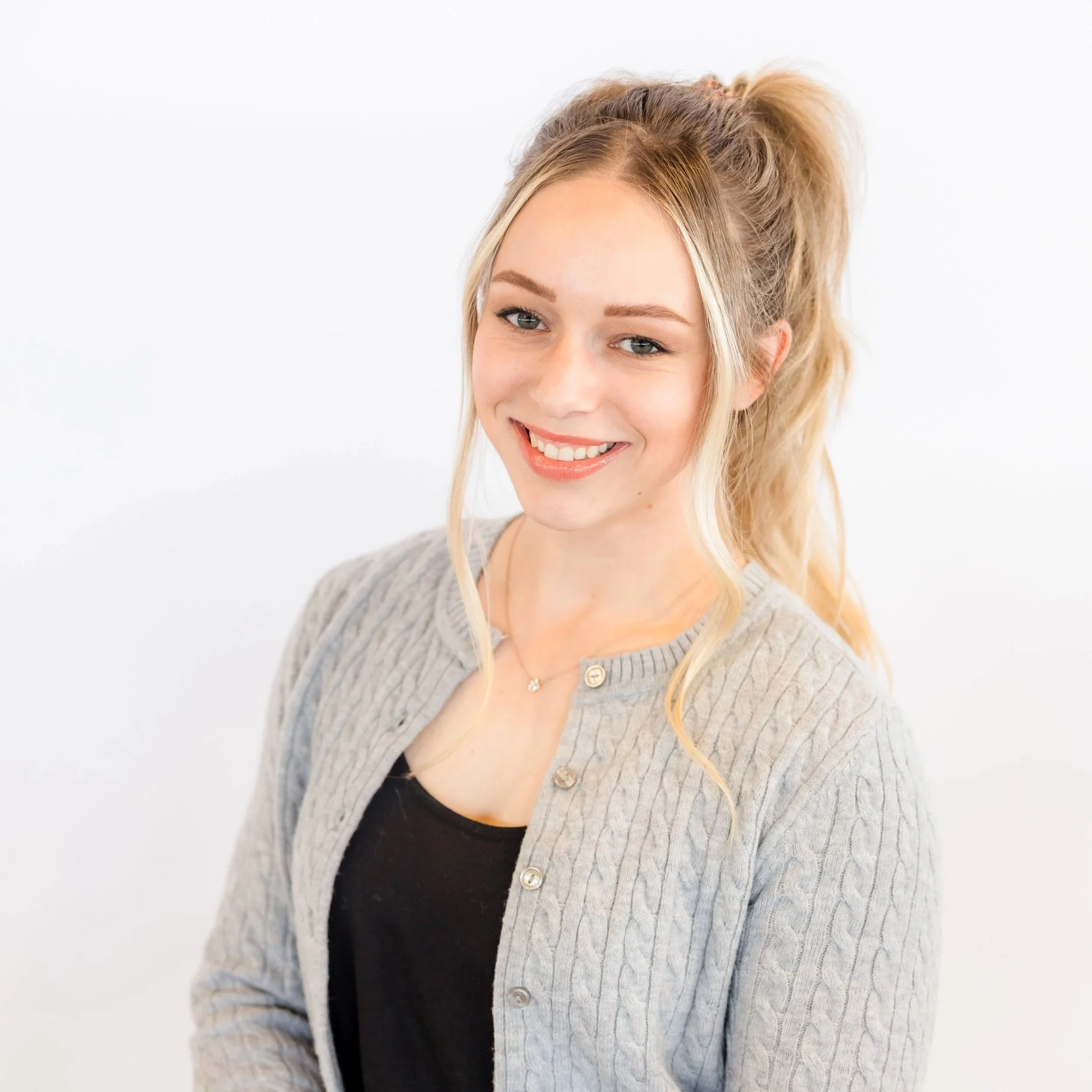 Close-up of a smiling woman with blonde hair tied in a high ponytail, wearing a grey knitted cardigan and a black top, standing against a white background.
