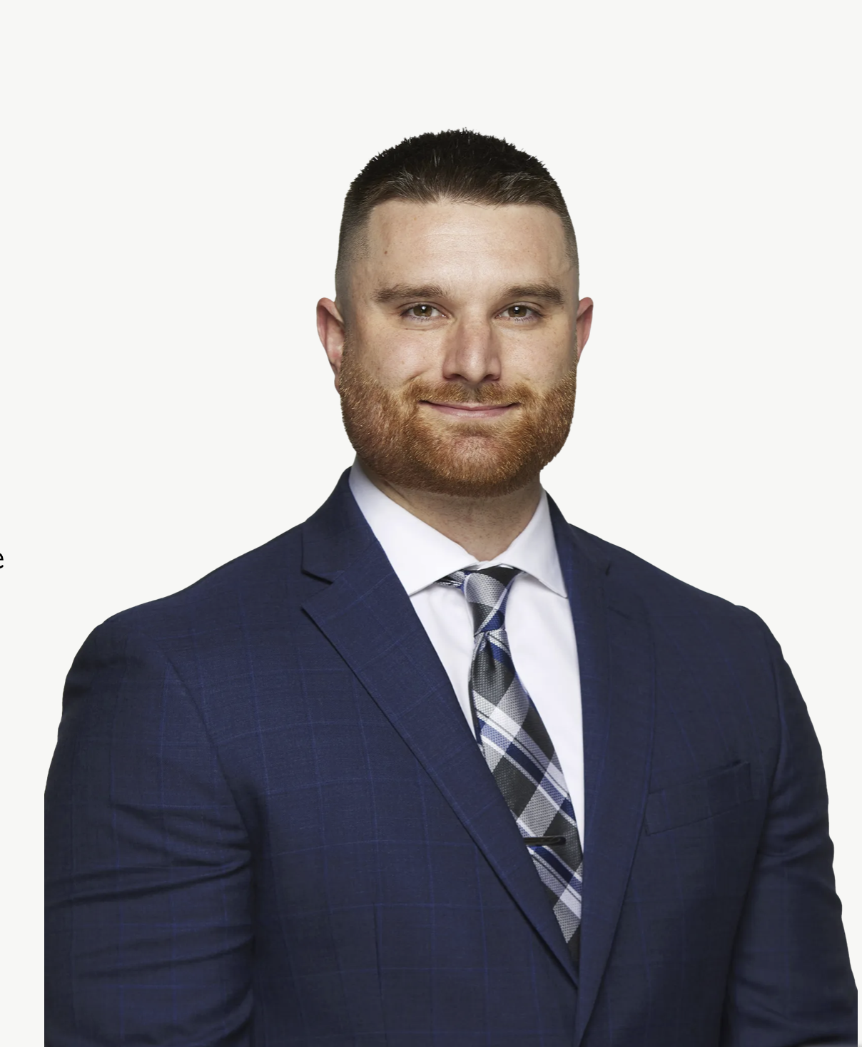 A man with short dark hair and a red beard wearing a dark blue suit, white shirt, and plaid tie, smiling at the camera.