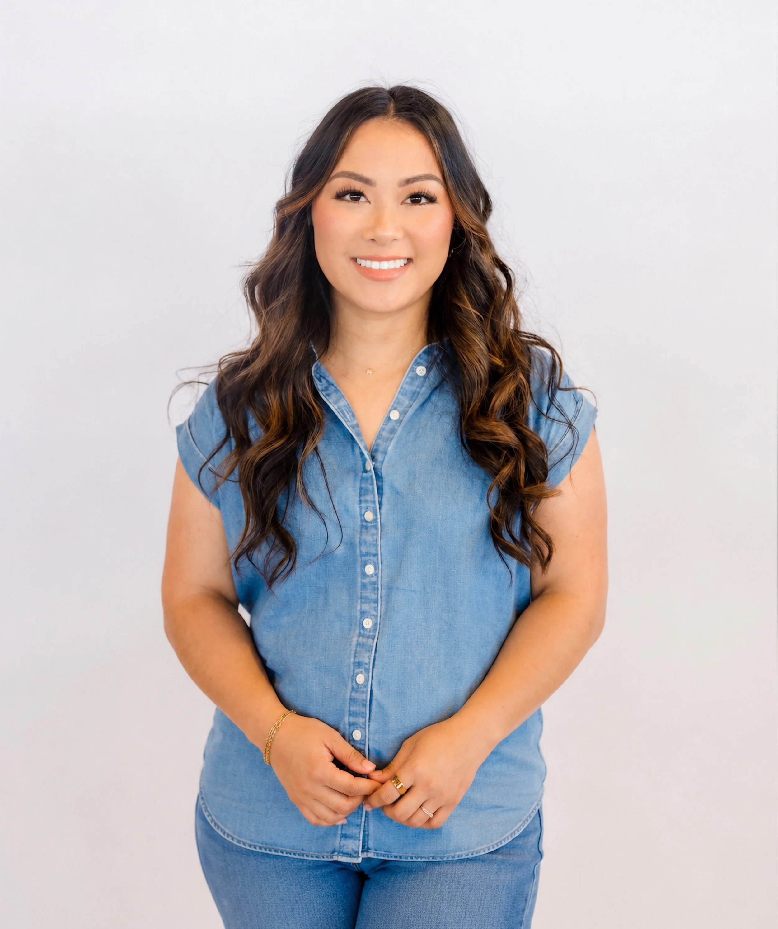 A woman with long wavy brown hair wearing a light blue denim shirt and matching jeans, standing in front of a plain white background, smiling with her hands clasped in front of her.