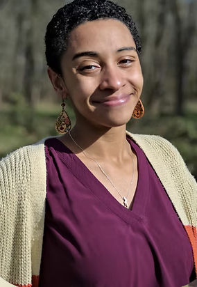 A woman with short, curly hair wearing a purple top, beige cardigan, and large earrings, smiling outdoors with trees in the background.