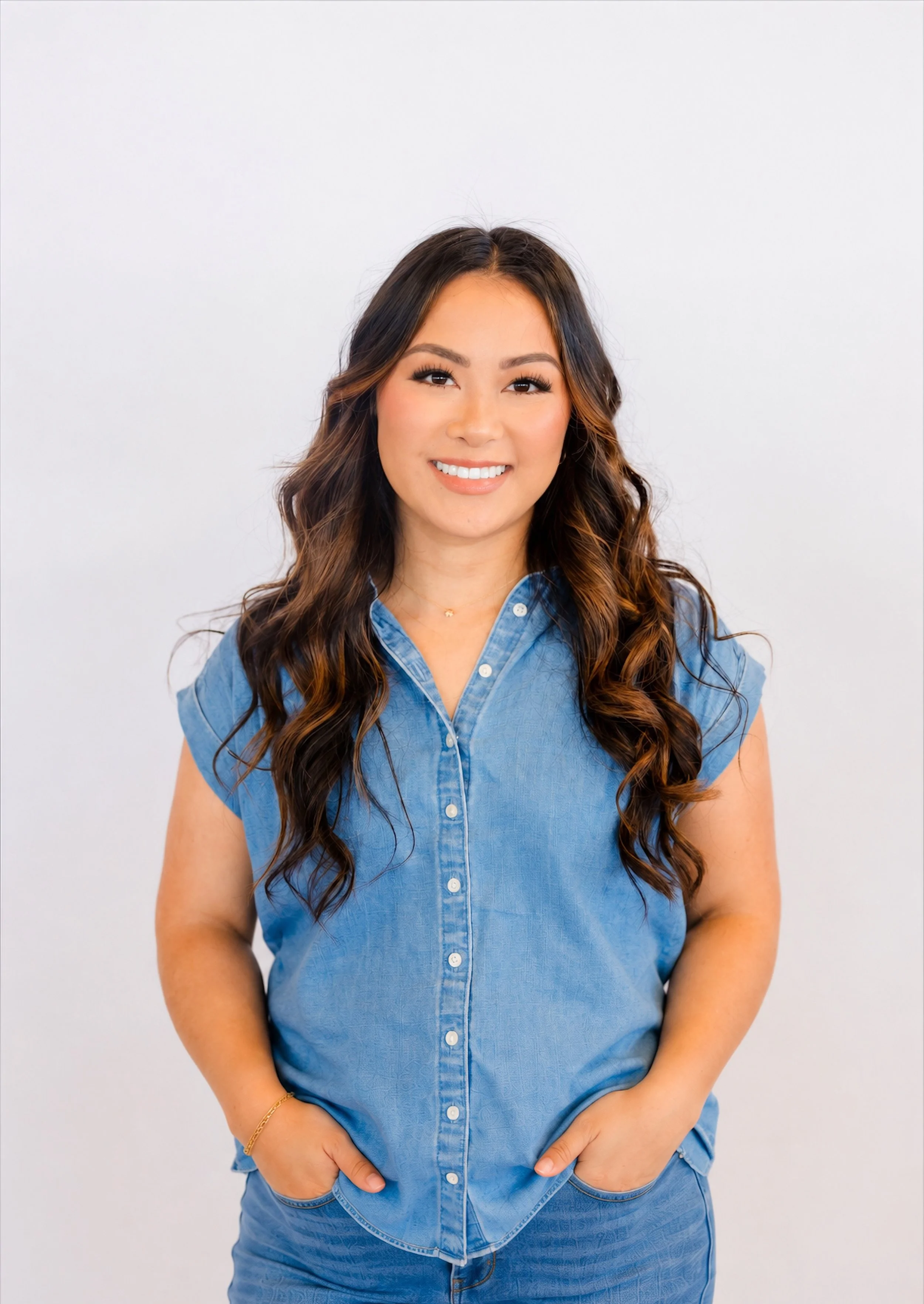A young woman with long, wavy brown hair, wearing a short-sleeved, button-up denim shirt and matching jeans, smiling and standing against a plain white background.