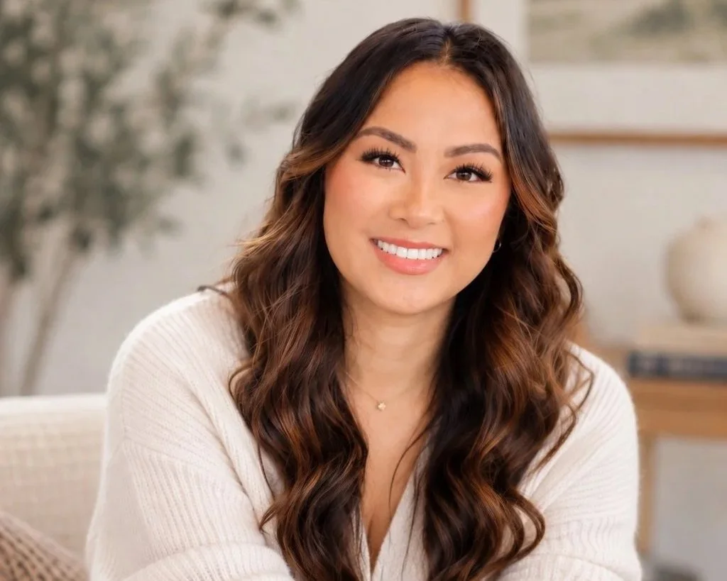A woman with long, wavy brown hair smiling at the camera in an indoor setting.