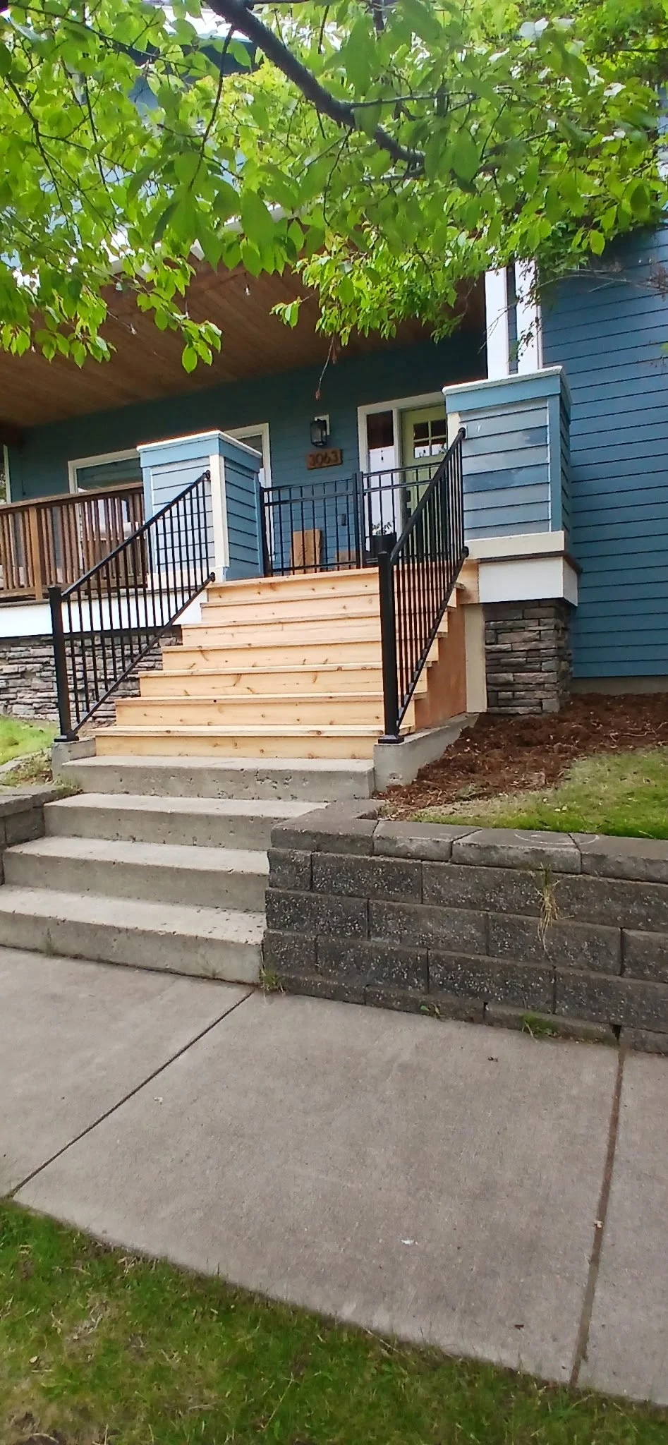 Front view of a house with new wooden stairs, black metal railings, a stone base, and a blue exterior with white trim under a tree.