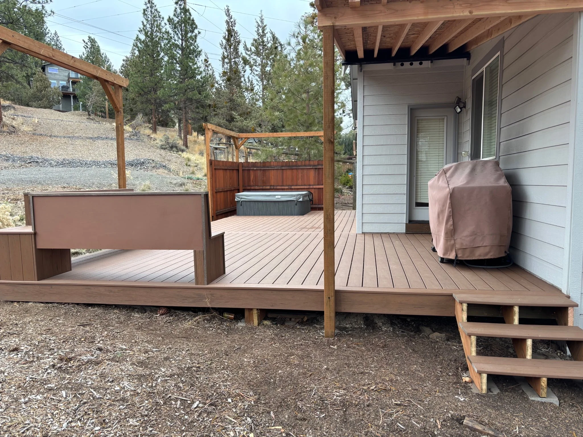 A backyard Trex deck with stairs leading down, surrounded by a wooden fence and tall pine trees in a hilly area. There is a covered hot tub on a concrete pad and outdoor lighting fixtures on the house wall. 