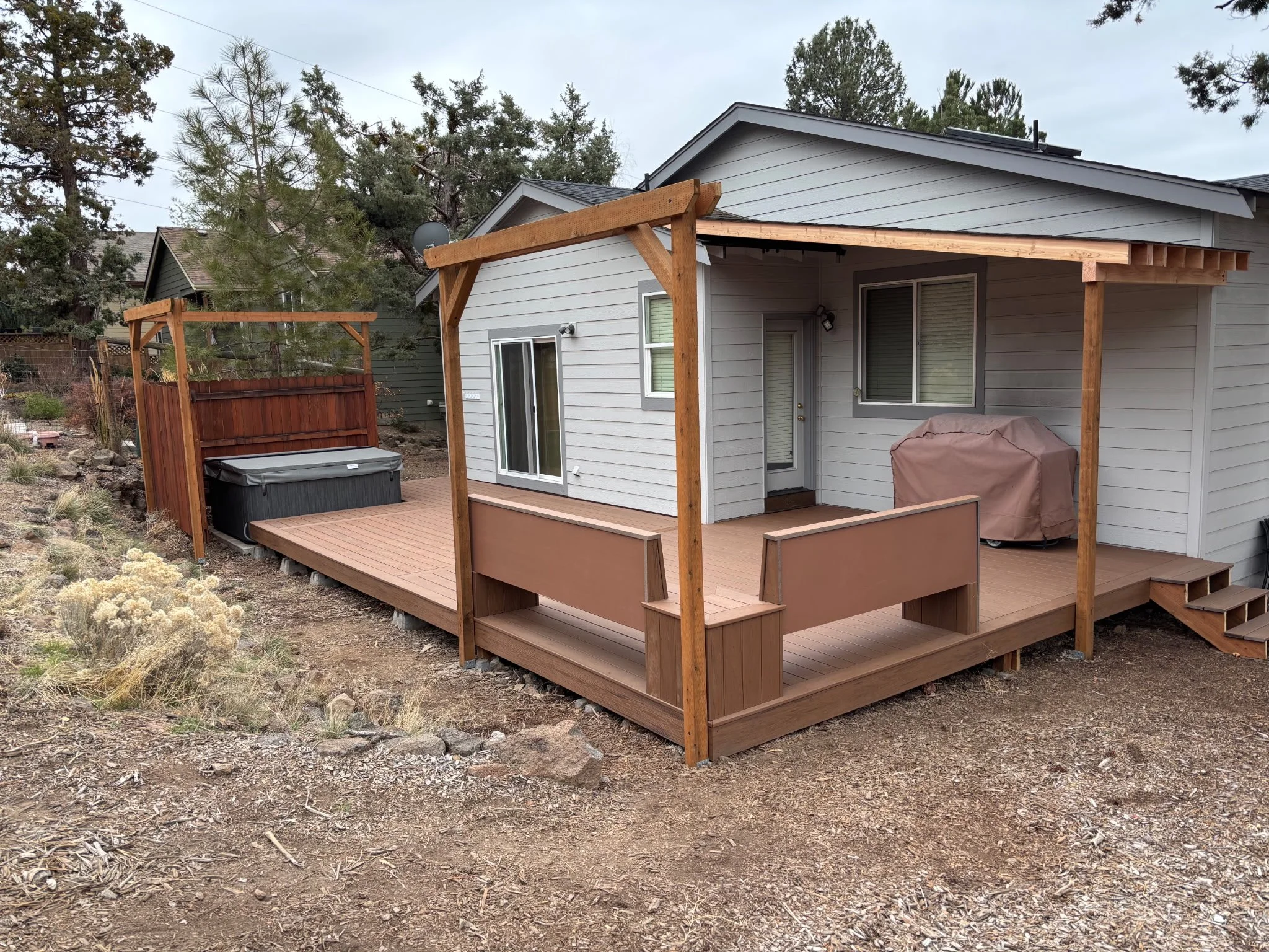 A backyard wooden Trex deck attached to a house with gray siding, featuring a pergola frame, a hot tub, and a covered grill, surrounded by dirt and plants with trees in the background.