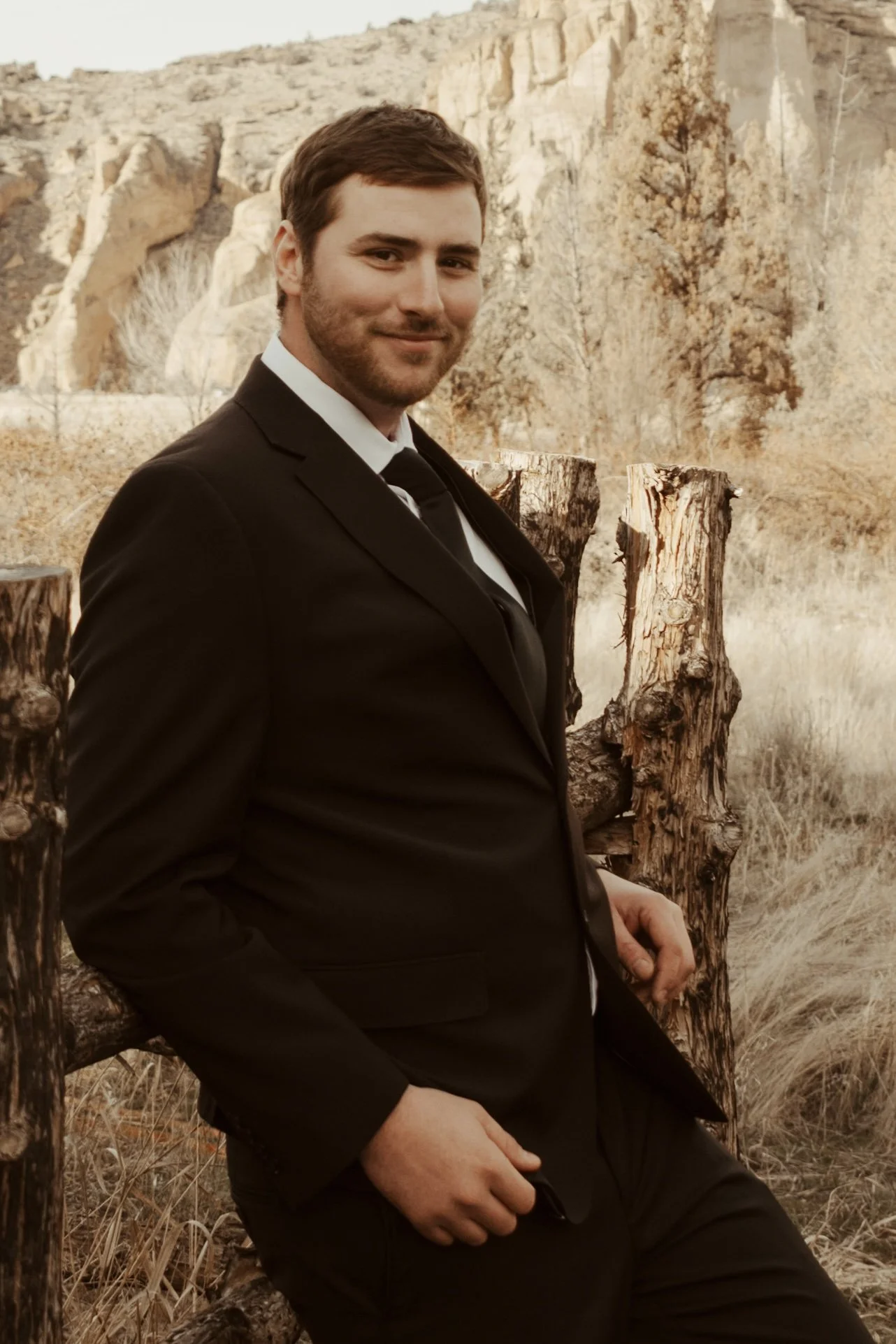 Man in a black suit sitting outdoors near wooden posts, with a desert landscape and rocky cliffs in the background.