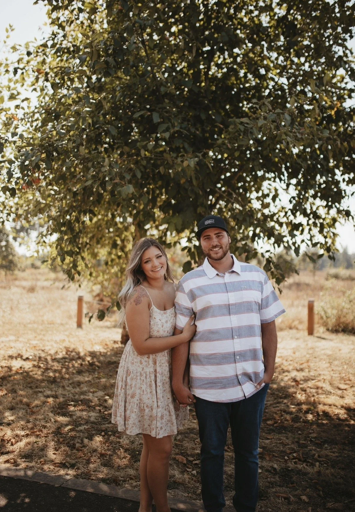 A couple standing outdoors under a large leafy tree, smiling at the camera. The woman has long, wavy hair and is wearing a floral dress with thin straps. The man has a beard, is wearing a baseball cap and a striped short-sleeve button-up shirt. The background shows a sunny day with dry grass and a wooden fence.