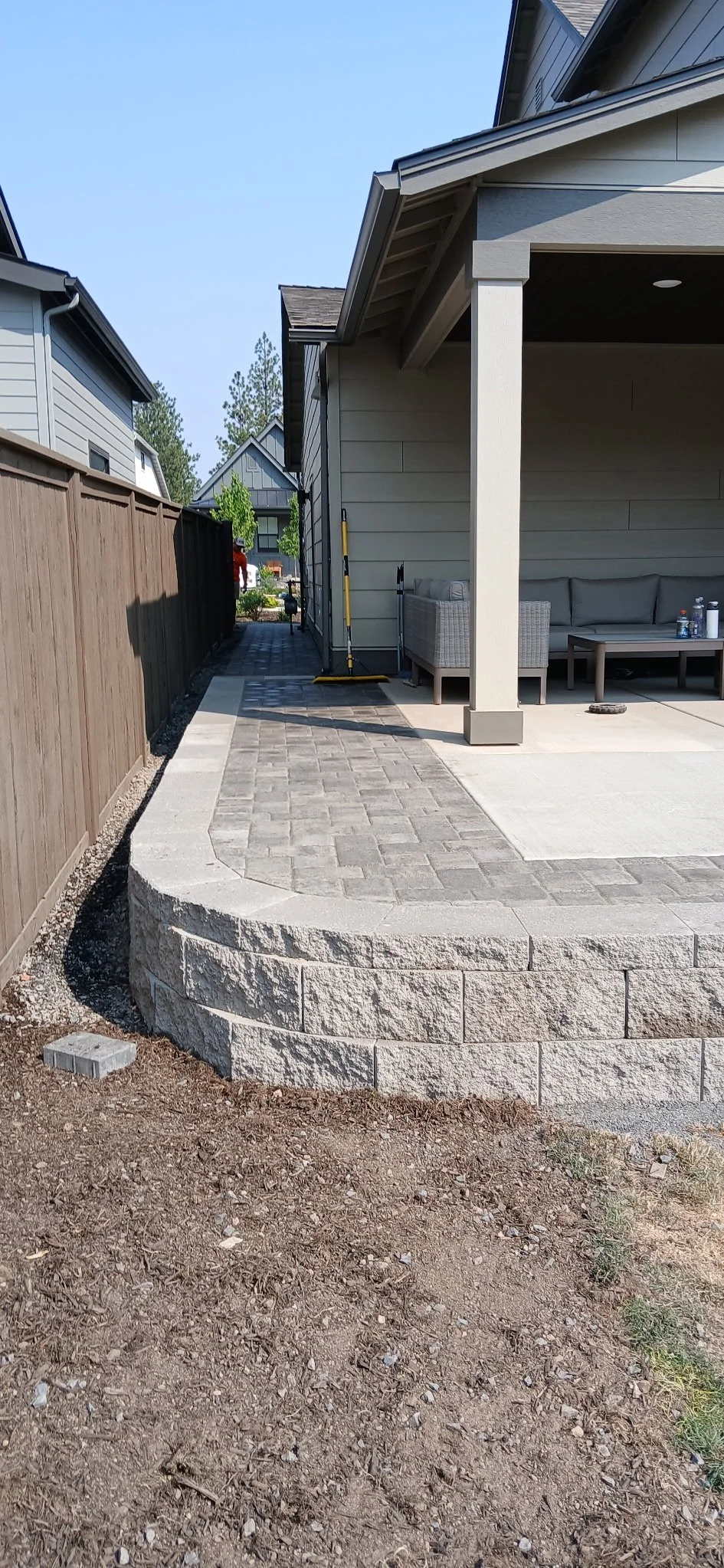 Back patio with paver patio, a stone retaining wall, and a wooden fence. There is outdoor furniture and construction tools on the porch, and dirt and soil in the foreground. Concrete hot tub pad.